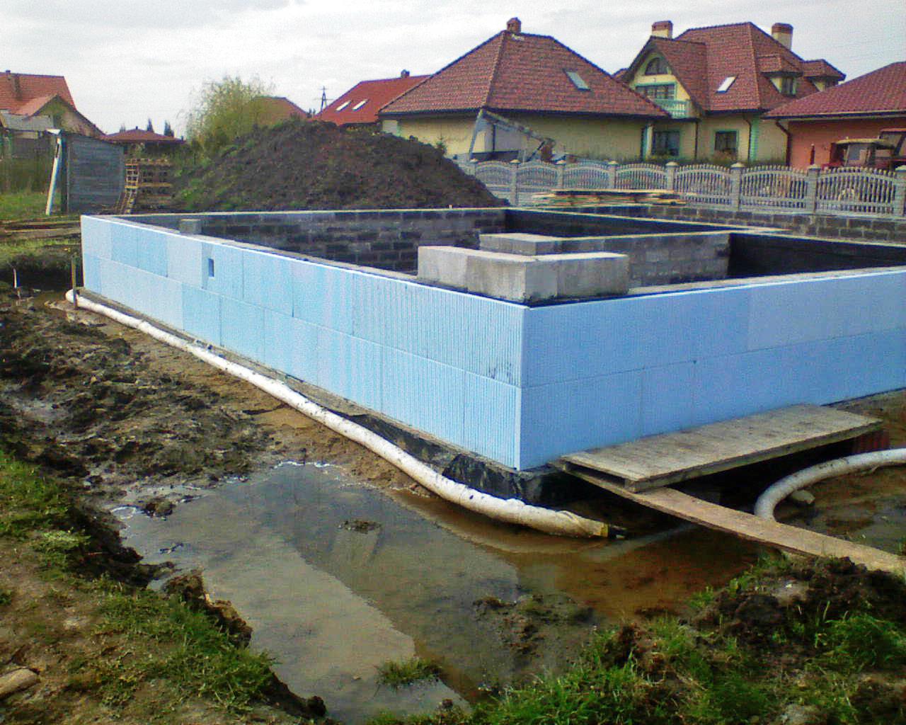 Niedokończona budowa fundamentów domu jednorodzinnego z widocznym szalunkiem, izolacją termiczną w postaci niebieskich płyt styropianowych, rurami drenażowymi oraz stertą ziemi w tle.